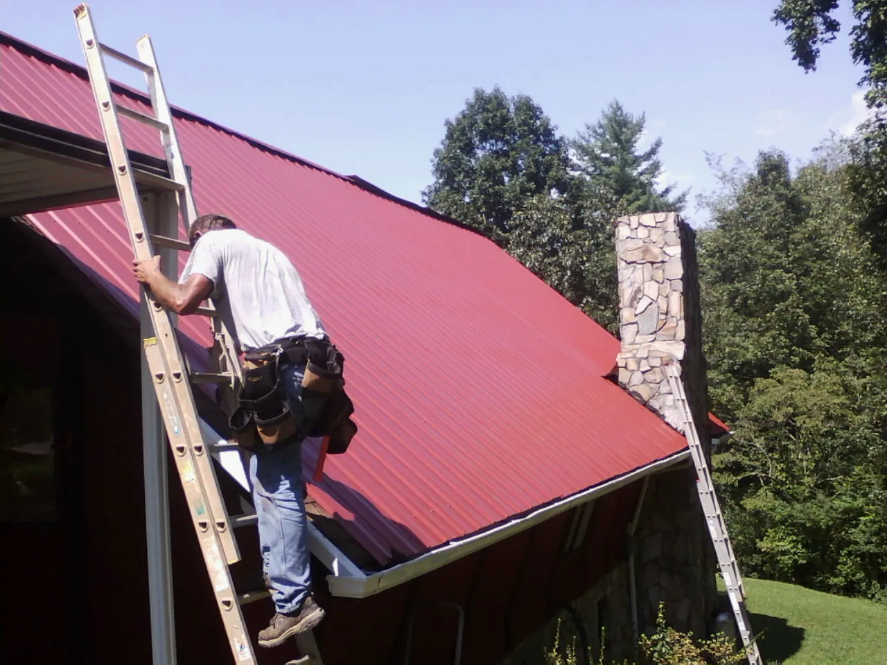 Roofer on a ladder beside a red metal roof and stone chimney — Soffit & Fascia Repair by Roofing Brothers in Willow Grove